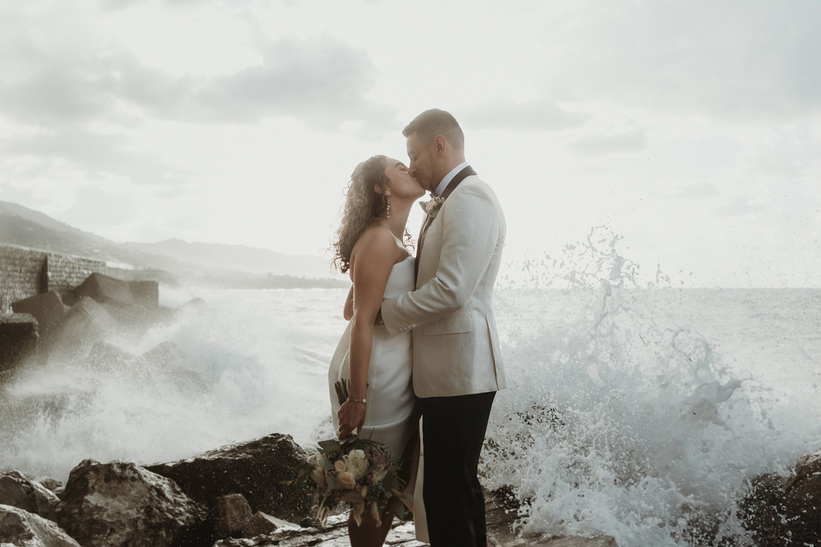 Couple kissing as a wave crashes on the rocks on their elopement in Cefalù, Sicily
