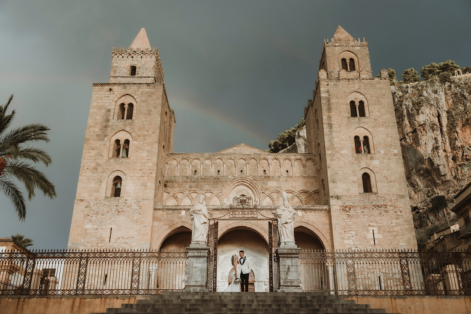 Couple standing in front of Duomo of Cefalù with dramatic sky and rainbow on their elopement in Sicily