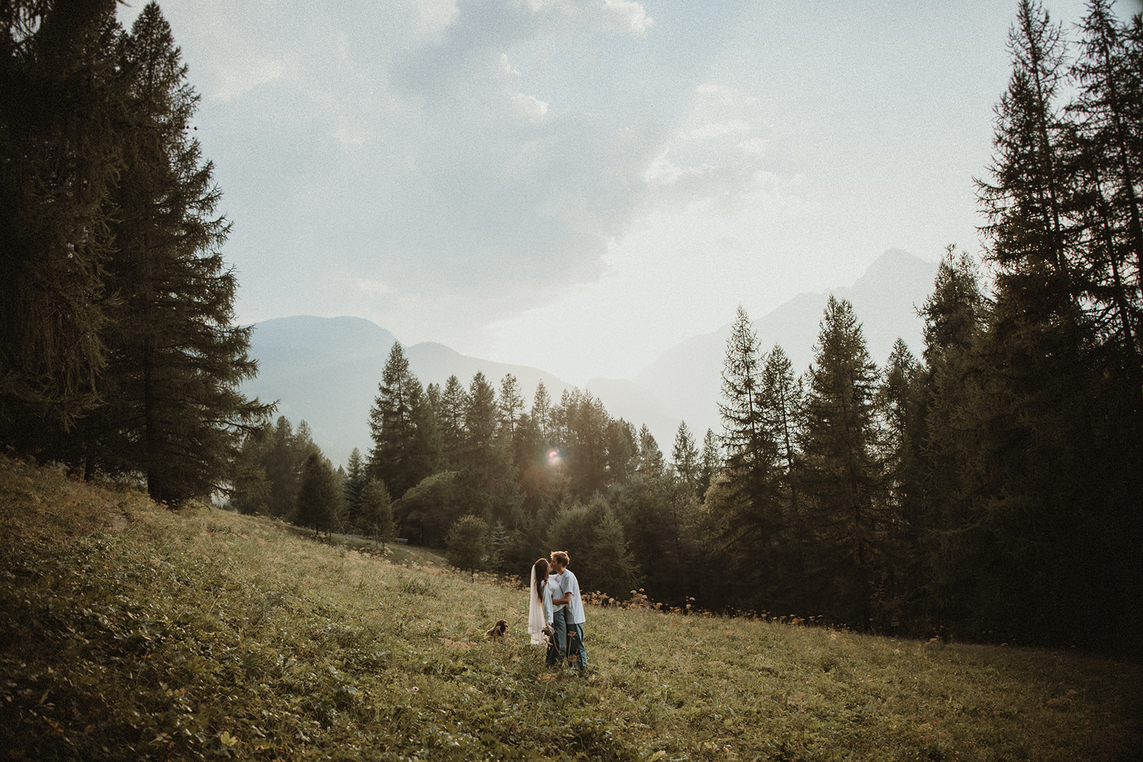 Couple kissing in a mountain meadow nestled in a forest with mountain views at the back on their Italian Alps Elopement