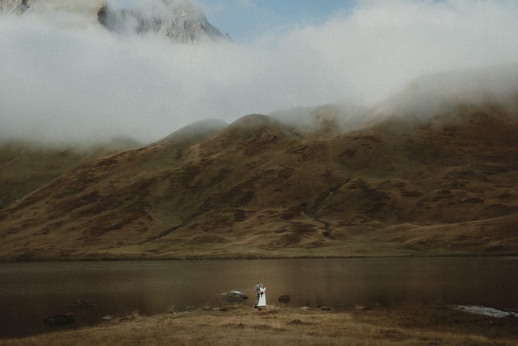 Couple kissing in the distance in a misty atmosphere with views of high altitude peaks and lakes in the Italian Alps on their elopement day