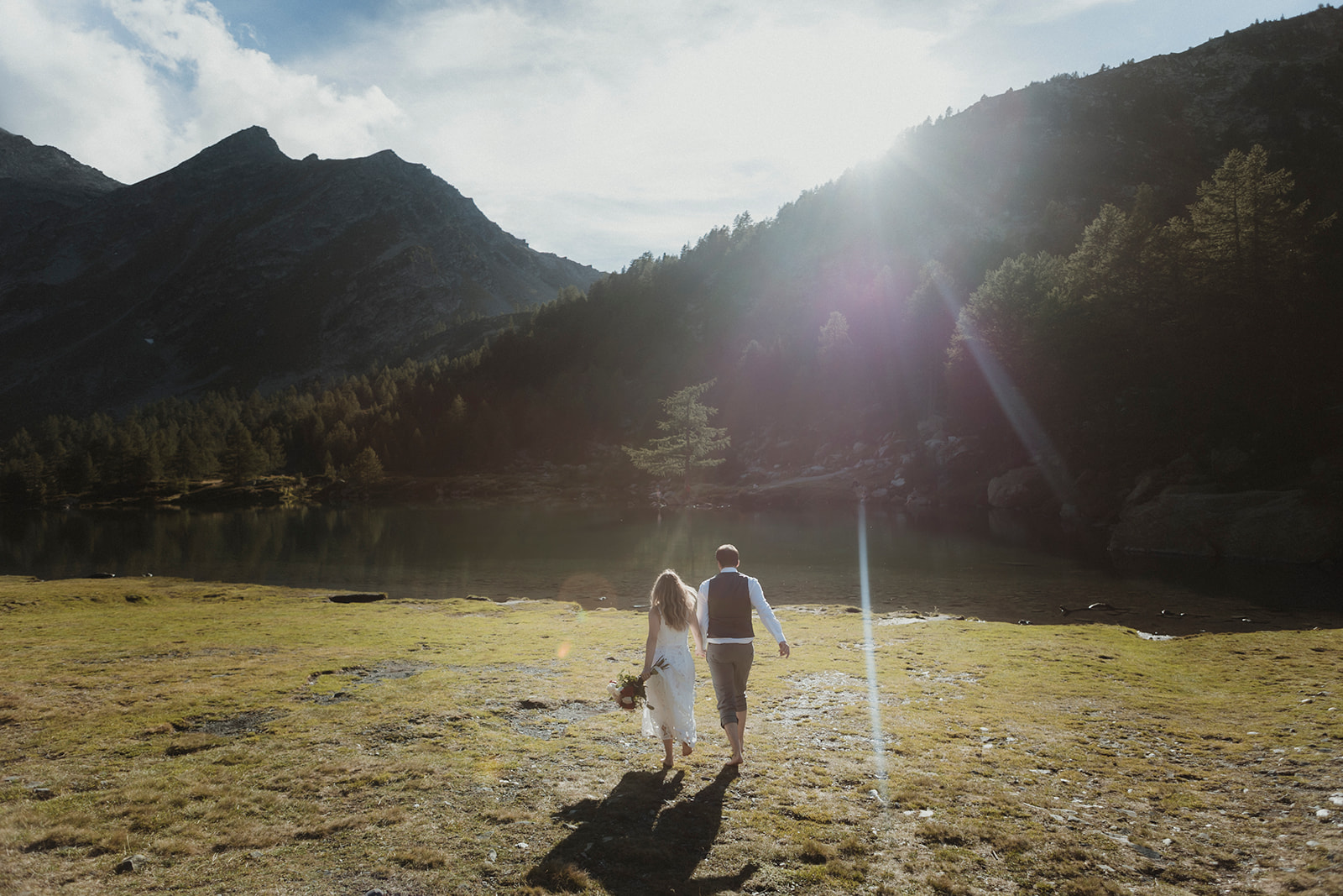 Couple walking in an alpine setting reaching their lake ceremony spot nestled within mountain peaks and forests Couple walking in an alpine setting reaching their lake ceremony spot nestled within mountain peaks and forests