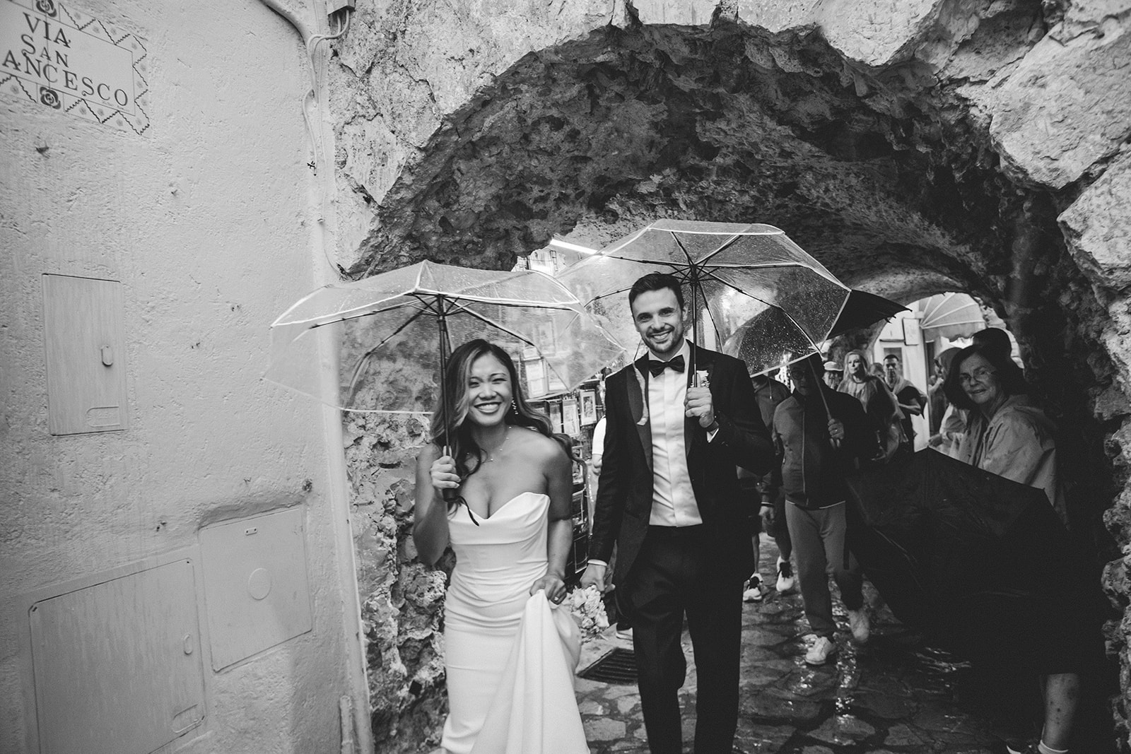 couple walking in the rain in a typical Italian town with their umbrellas on their elopement day
